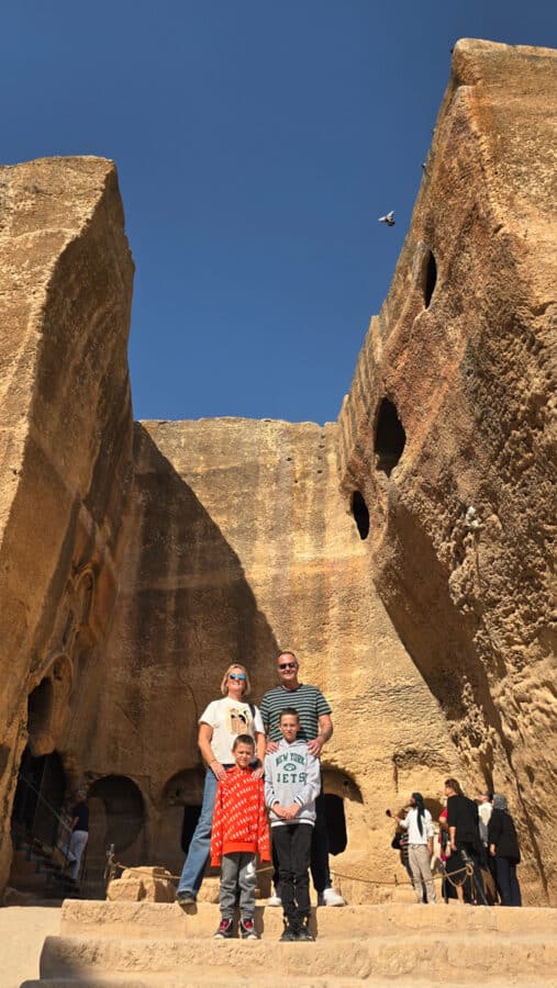 A group of people stands in front of the large ancient stone ruins of the Ancient City of Dara, with a clear blue sky and a bird flying overhead, evoking the rich Mesopotamian heritage of Mardin, Turkey.