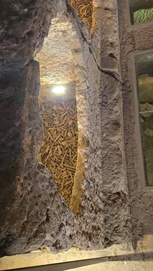 A narrow stone chamber filled with stacked skeletal remains, illuminated by an overhead light, reflects the rich Mesopotamian heritage of the ancient city of Dara in Mardin, Turkey.