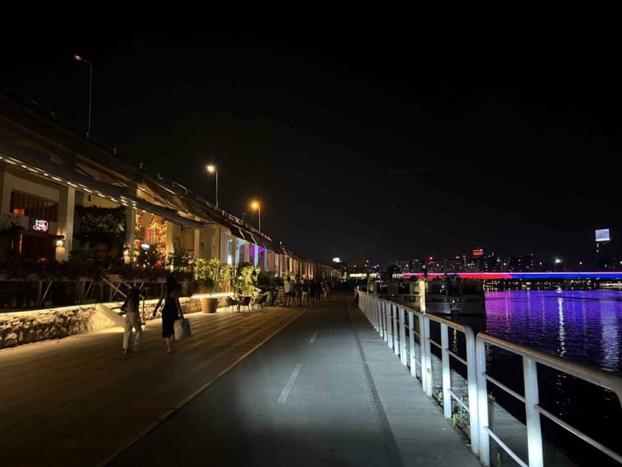 A nighttime scene of a riverside walkway in Belgrade, where people stroll alongside lit buildings on the left, embraced by the vibrant energy of Belgrade nightlife, with colorful reflections dancing on the water to the right.