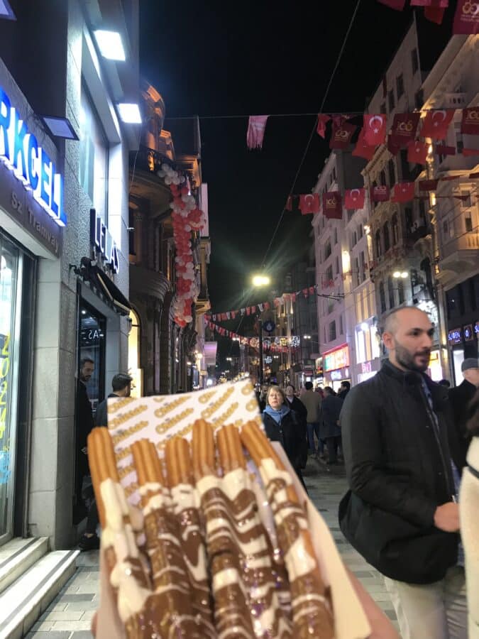 A person holds a box of churros drizzled with chocolate on a busy, flag-decorated street in Istanbul at night. People savored the festive atmosphere, reminiscent of Christmas, as they strolled past charming shops lining the bustling pathway.