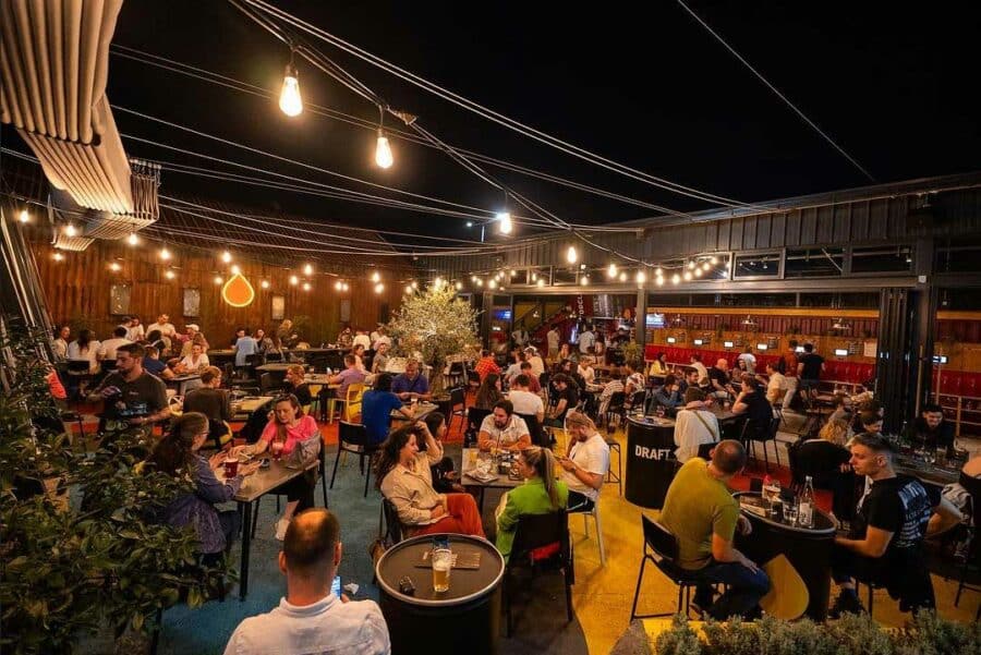 A lively outdoor bar scene at night in the heart of Belgrade, with numerous people seated under string lights, engaging in conversations and enjoying drinks. It's the epitome of Belgrade nightlife—one of the best places to party and soak up the city's vibrant energy.