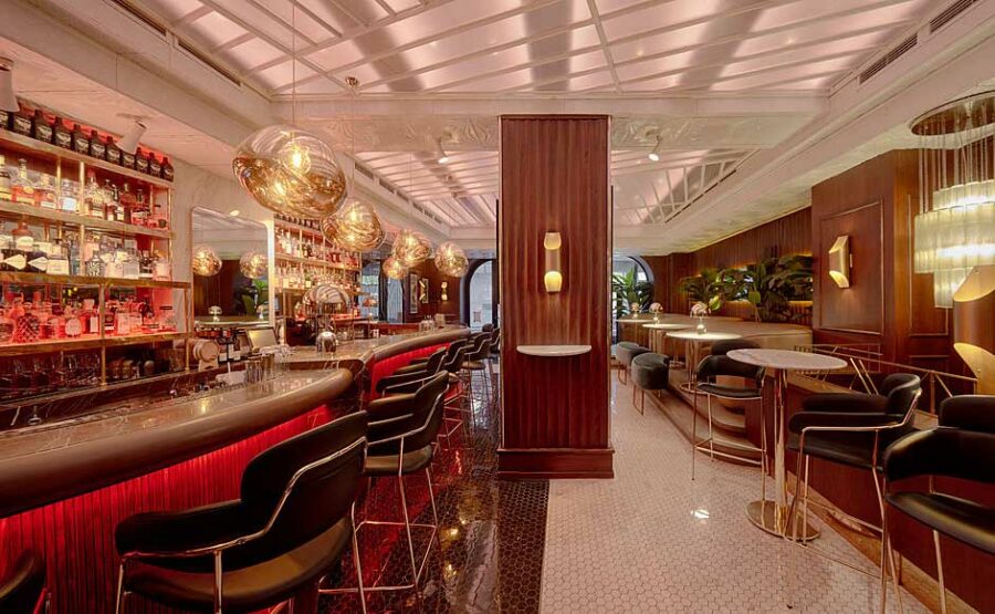 Modern bar interior with a curved bar counter, red lighting, and round hanging lamps captures the vibe of Belgrade nightlife. Rows of bottles are displayed on shelves, while tables and chairs are carefully arranged along a tiled floor, making it one of the top places to party.