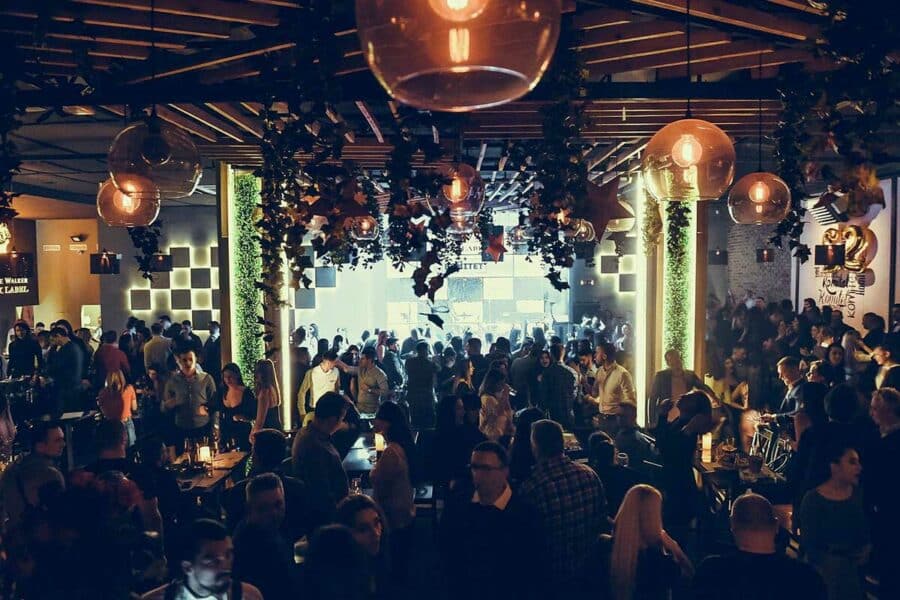 A lively Belgrade nightlife scene with people dancing and socializing under dim lighting and decorative hanging lights captures the essence of the party atmosphere.