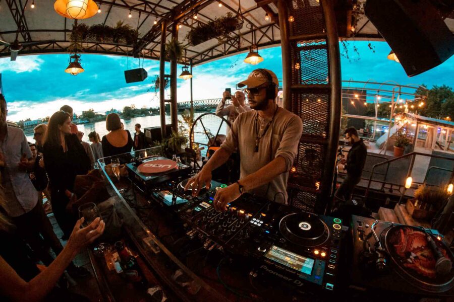 A DJ spins tunes at an outdoor venue, igniting the Belgrade nightlife. The vibrant party scene unfolds with a lively crowd, state-of-the-art equipment, and a stunning river view under the enchanting evening sky.