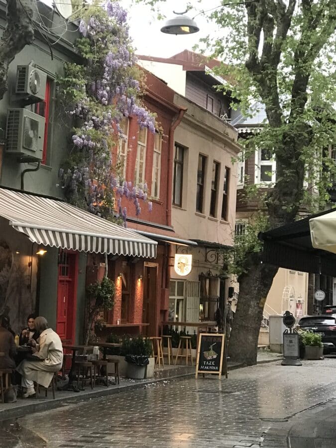 A quaint street in Kuzguncuk with colorful buildings, outdoor seating at a café, lush wisteria, and a wet cobblestone path. Two people are seated, enjoying the serene vibe as a small chalkboard sign guides visitors to this charming spot.