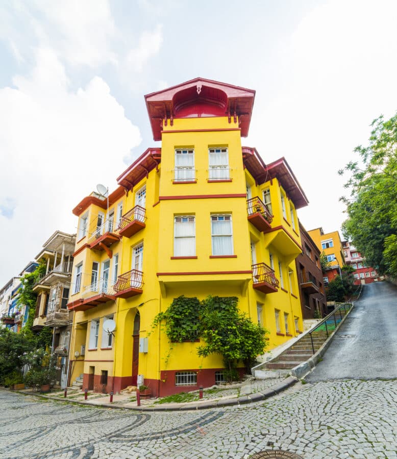 In the heart of Kuzguncuk, a vibrant yellow multi-story building with red trim and balconies graces a cobblestone street corner. Surrounded by other colorful buildings and lush greenery, this picturesque sight is like stepping into a guidebook illustration.