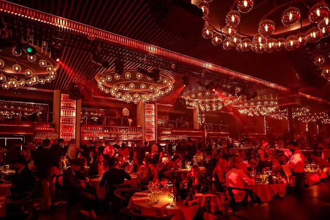 A dimly lit, bustling bar with red lighting and chandeliers captures the essence of Belgrade nightlife, as patrons revel in vibrant conversations at their tables.