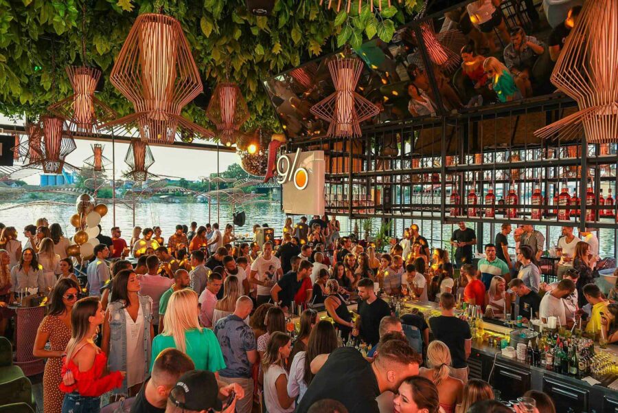 A crowded bar buzzes with people socializing under hanging lights, enveloped by lush plants and a stunning view of the water—an ideal spot to dive into the vibrant Belgrade nightlife.