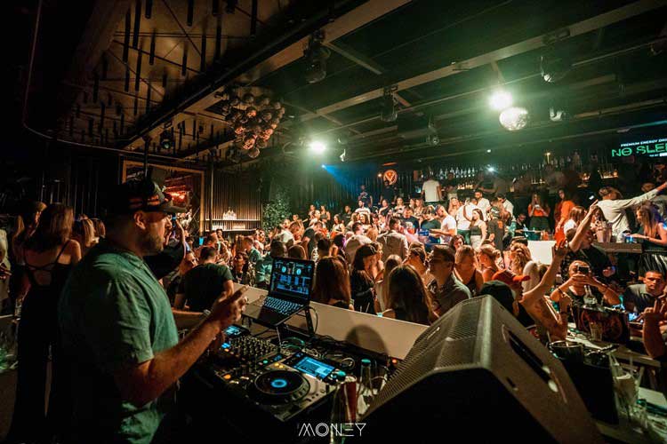 A DJ is performing in a crowded nightclub, capturing the essence of Belgrade nightlife, with people dancing and socializing. Sound equipment is visible in the foreground as the party vibe electrifies the room.