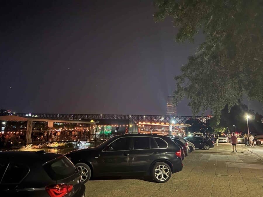 The night scene showcases Belgrade's vibrant nightlife, with parked cars in the foreground and a crowded outdoor area bustling on the left. A lit bridge glows under the dark sky, inviting visitors to explore one of the city's top places to party.