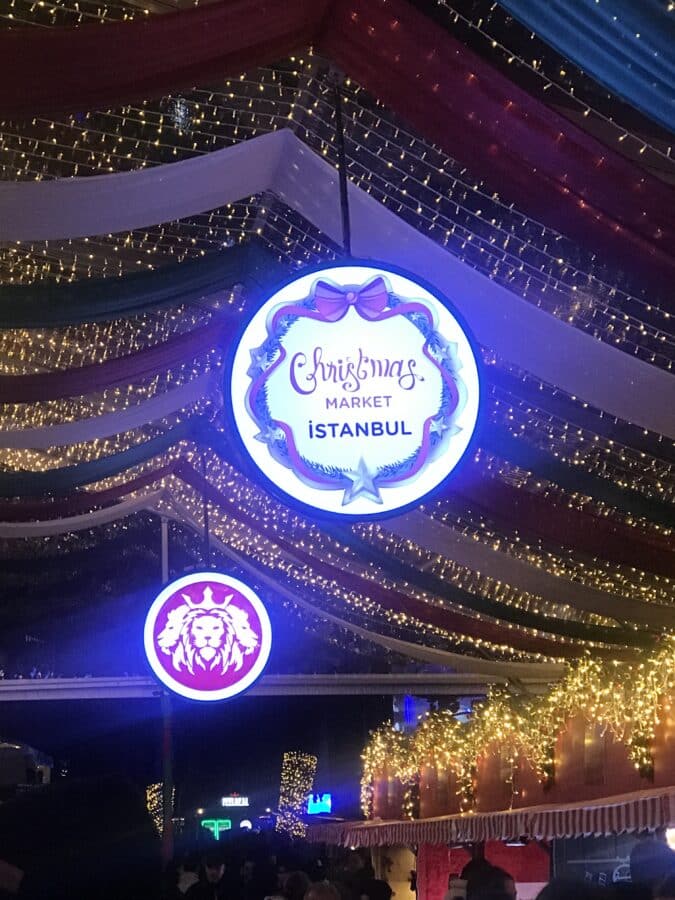 Illuminated signs and festive lights adorn the Christmas market in Istanbul at night, inviting visitors to spend time exploring beneath a circular sign proudly displaying "Christmas Market Istanbul.