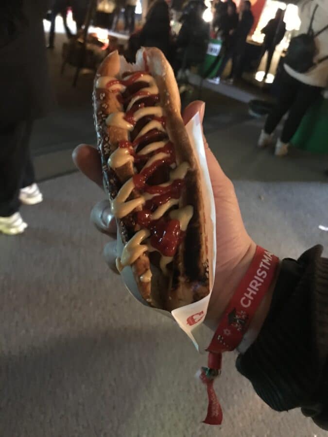 Amidst the lively stalls and bustling crowd, a person enjoys a hot dog smothered in mustard and ketchup. The scene feels almost festive, reminiscent of an Istanbul street bazaar on Christmas, alive with sights and sounds.