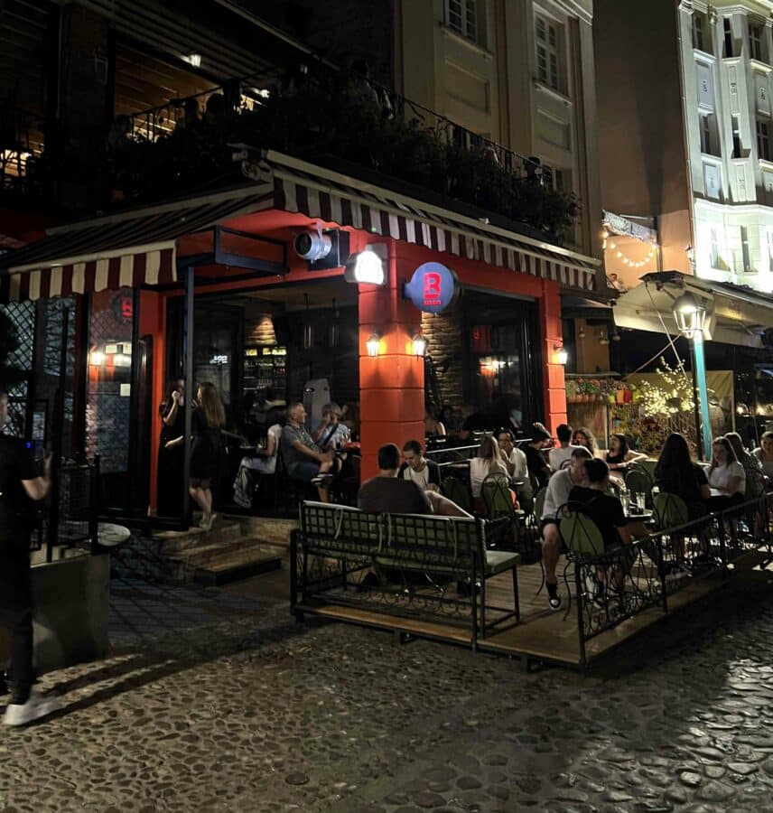 People sit and converse outside a cozy, dimly lit café with red accents on a cobblestone street at night, capturing the essence of Belgrade nightlife.