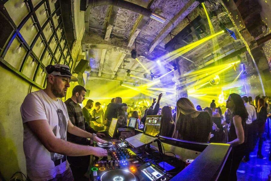 A DJ wearing headphones performs at a lively nightclub in Belgrade, surrounded by people dancing. Colorful lights and a disco ball illuminate the vibrant scene, capturing the essence of Belgrade nightlife.