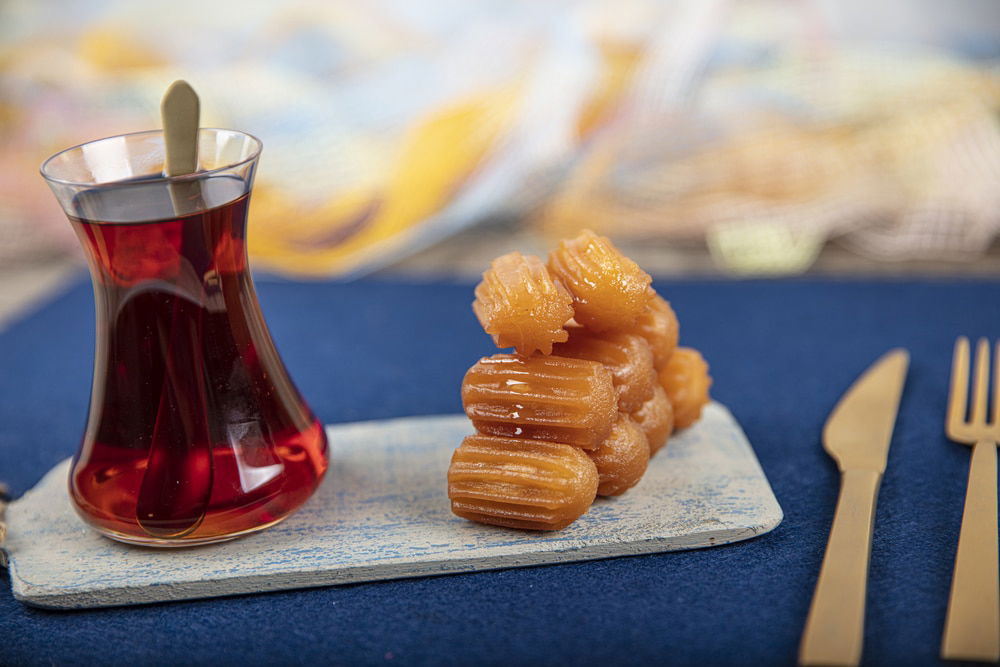 A glass of tea and a stack of syrupy pastries on a small wooden board, with gold-colored cutlery on a blue surface.