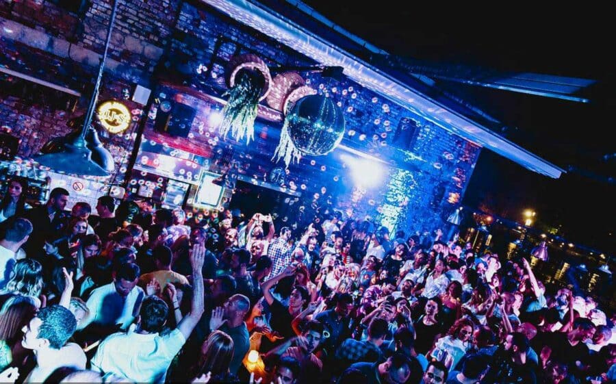 A lively indoor party with a large crowd dancing under colorful lights and disco balls captures the essence of Belgrade nightlife.