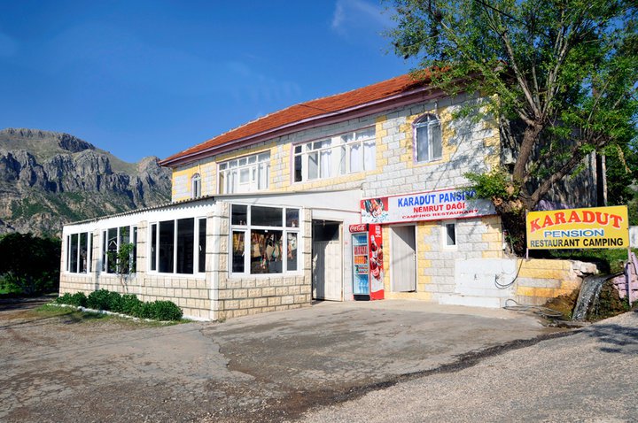 A stone building with a sign for Karadut Pension Restaurant Camping stands proudly against the majestic backdrop of Mount Nemrut, framed by clear skies.