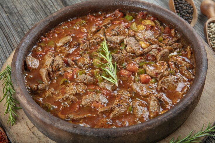 A clay dish filled with a meat stew, garnished with a sprig of rosemary, sits on a wooden surface.