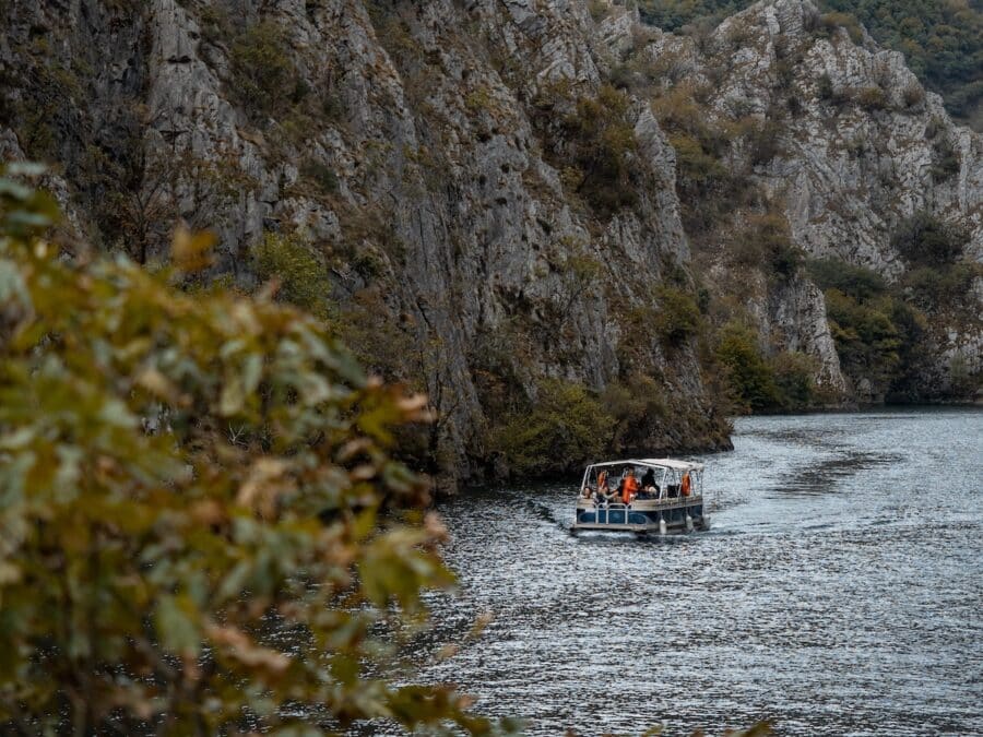 A small boat with people, some wearing orange life vests, sails through the serene waters of Matka Canyon, surrounded by rocky cliffs and dense greenery.