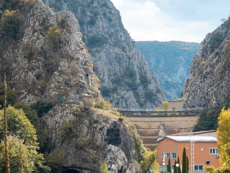 A hydroelectric dam nestled between the rocky cliffs of Matka Canyon, near Skopje, stands proudly with a building in the foreground and lush greenery surrounding the structure.