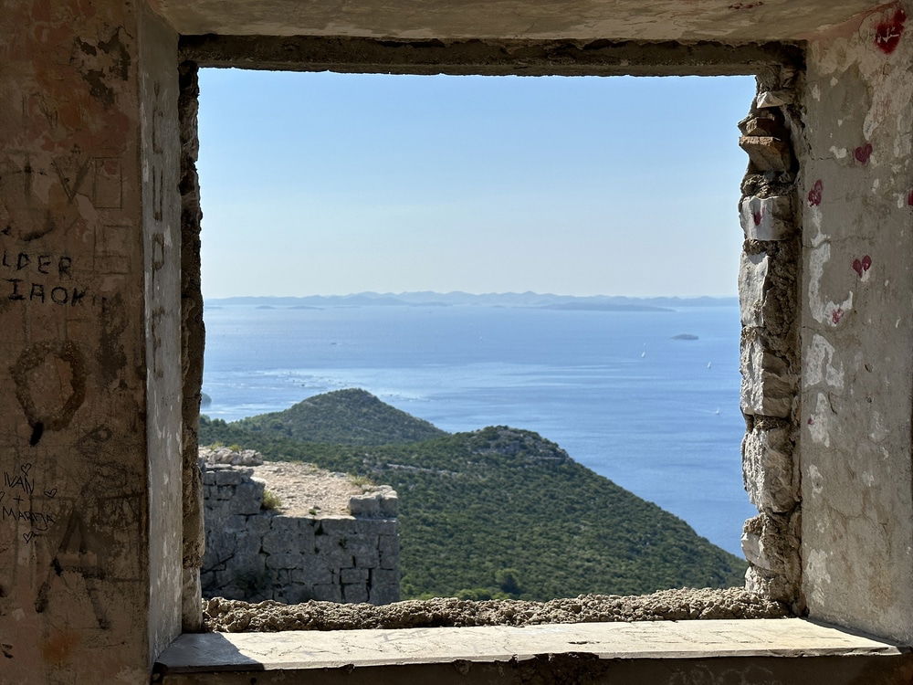 Through the stone window, discover the serene landscape of green hills gently sloping toward the vast blue ocean under a clear sky, with islands like Ugljan and Pašman dotting the horizon near enchanting Zadar.