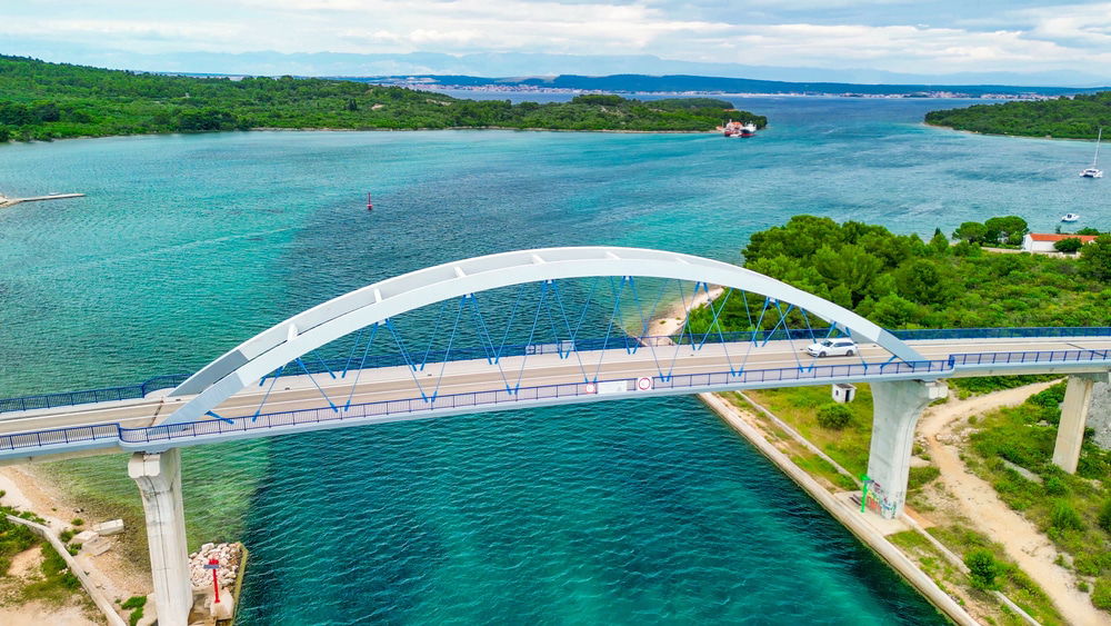 Aerial view of a white arch bridge spanning the turquoise waters near Zadar, with a car crossing it, surrounded by the lush green landscape of Ugljan and distant islands like Pašman.