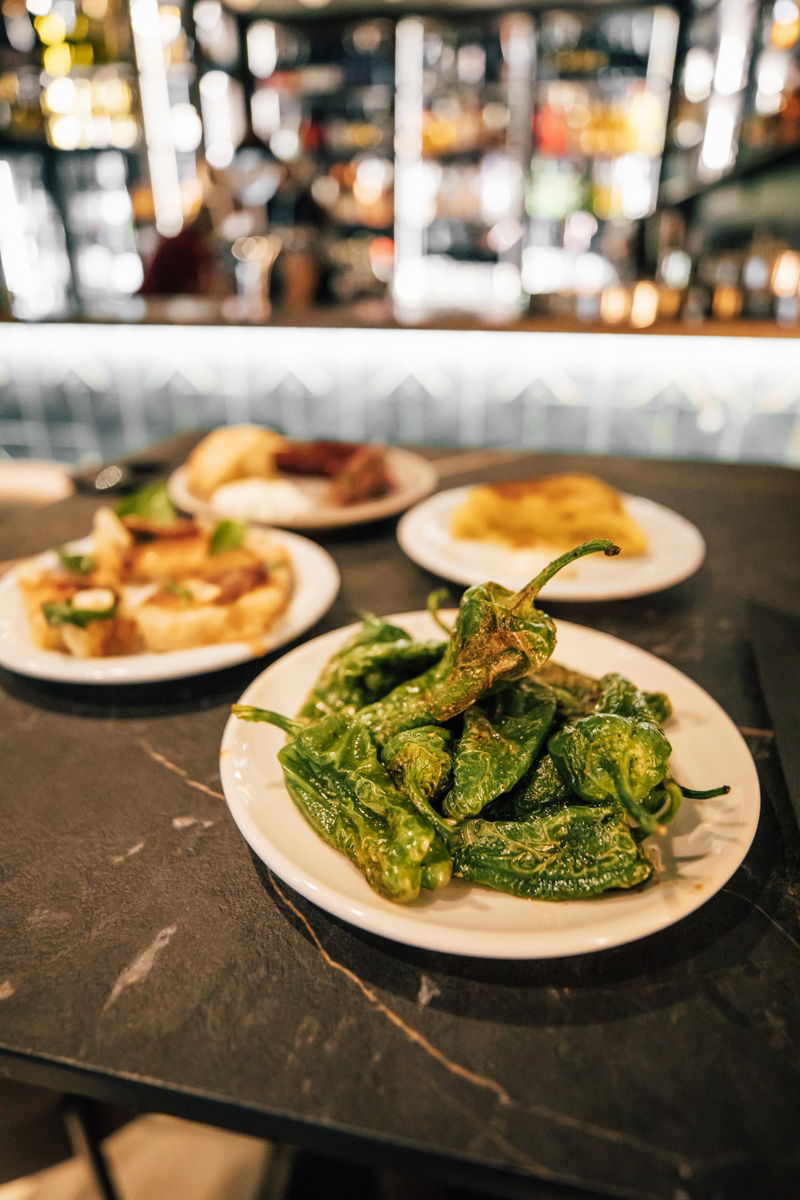 A plate of blistered green peppers takes center stage on a dark table at Pinčo Tapas & Wine Bar in Zadar, with three plates of assorted tapas softly blurred in the background.