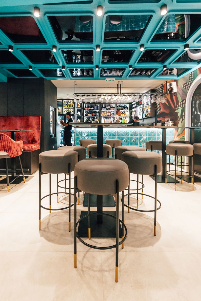Modern wine bar interior with high stools, a patterned blue ceiling, illuminated bar counter, and shelves stocked with bottles; the bartender is working behind the counter at Pinčo Tapas & Wine Bar in Zadar.