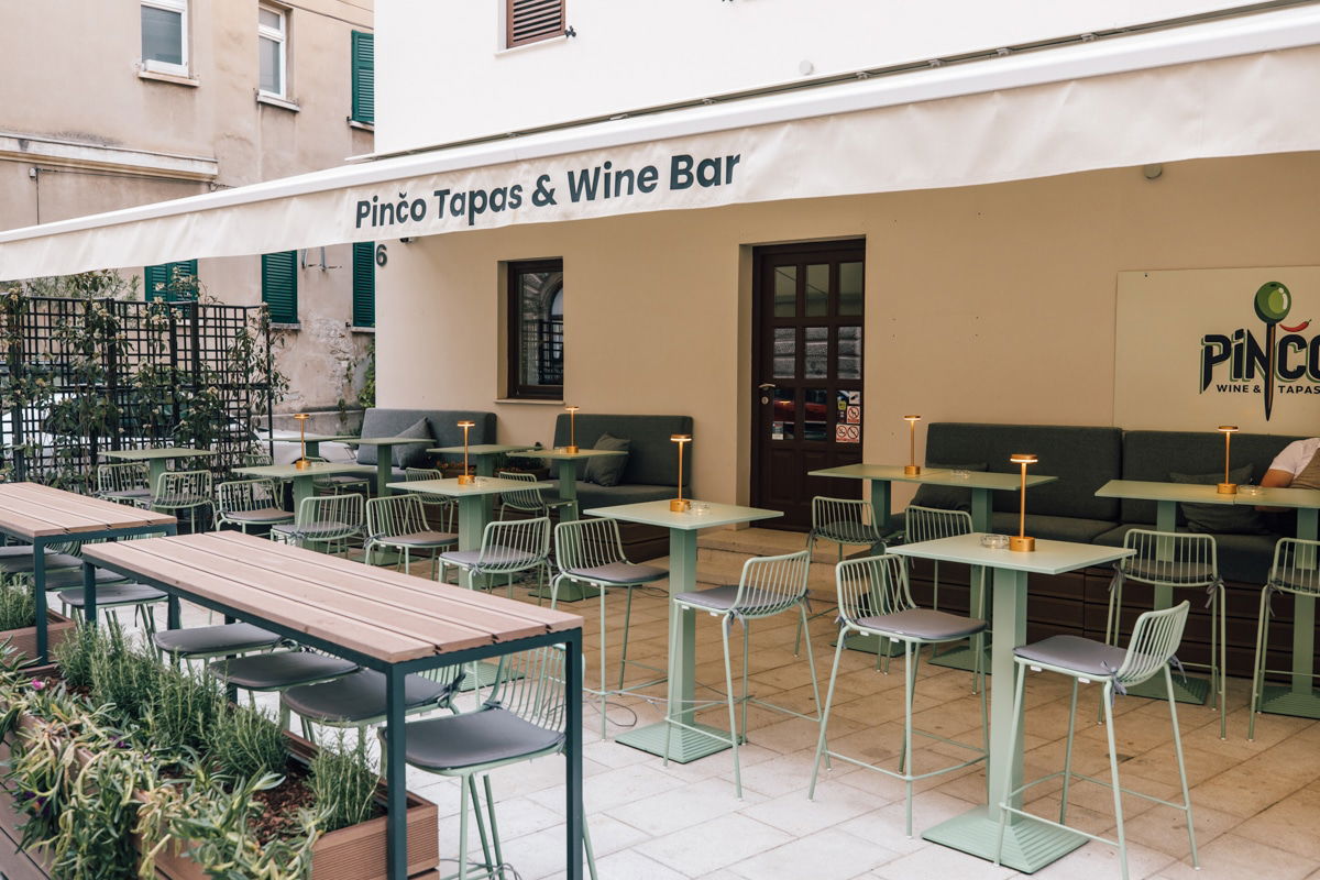 Outdoor seating area of Pinço Tapas & Wine Bar in Zadar, featuring light green tables and chairs, cushioned benches, small table lamps, and potted plants on a tiled patio.