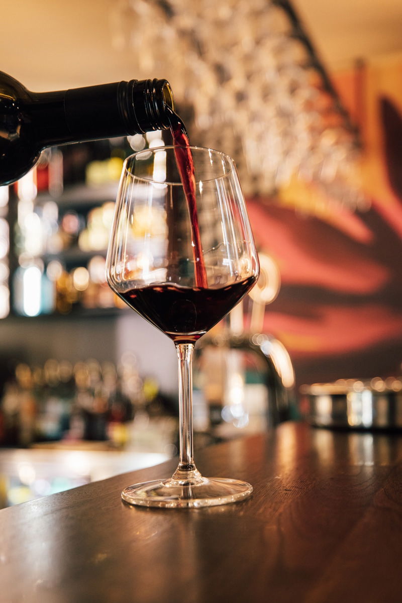 Red wine being poured from a bottle into a wine glass on a bar counter at Pinčo Tapas, with shelves of bottles blurred in the background at this cozy Wine Bar Zadar.